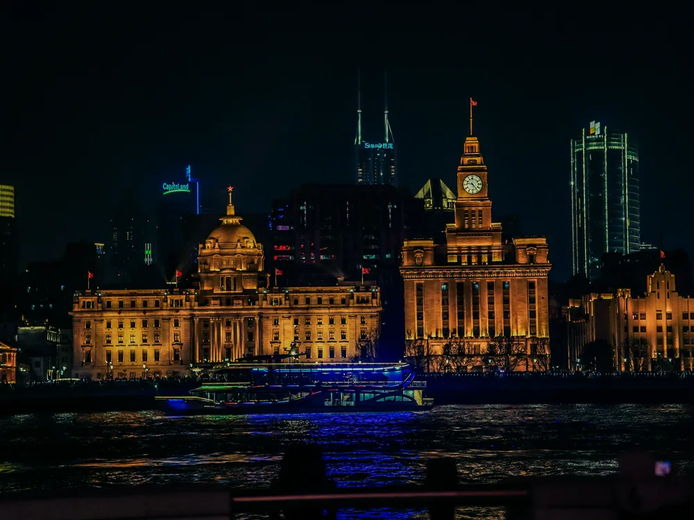 A cruise boat lit blue on the river in front of the Bund's gold-lit historic buildings and clock tower at night
